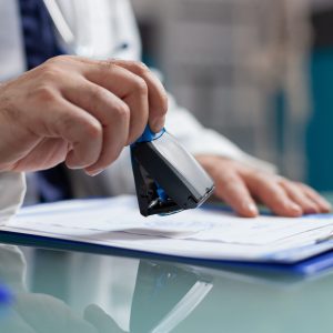 Doctor holding medical stamp to put on prescription paper at health care exam. Hand of physician putting seal on checkup report, giving medicine to patient at consultation. Close up